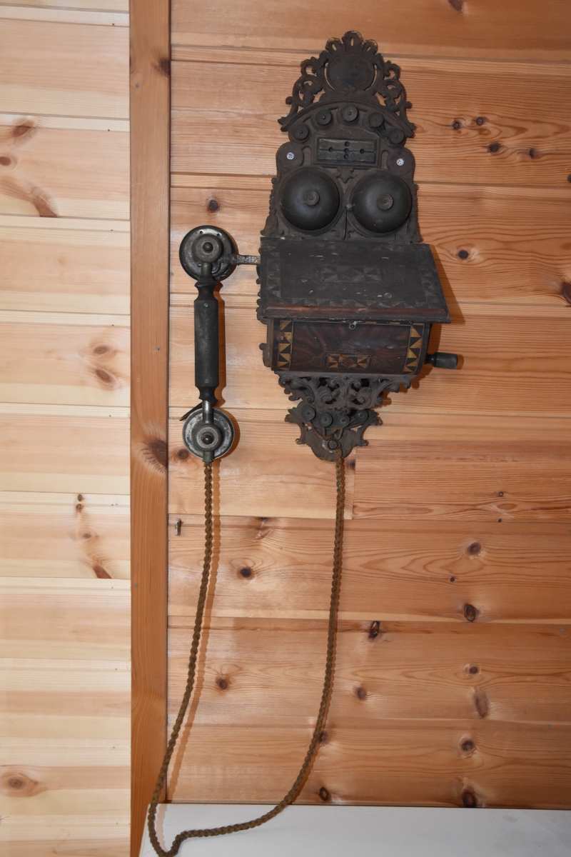 Magneto veggtelefon av blikk med ornamentert støpejern. Treimitasjon og Geometriske dekorasjoner på lokk og fremside som kan minne om treimitasjon. Stoffledning som er tvunnet. Tekst i øvre del. Sveiv på høyre side. Røret med høytaler og"taltetut" henger på venstre side.