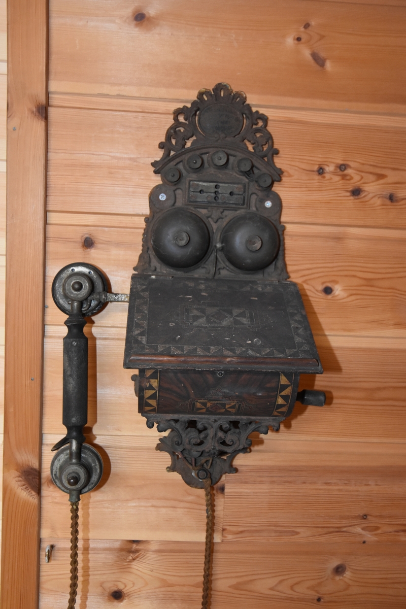 Magneto veggtelefon av blikk med ornamentert støpejern. Treimitasjon og Geometriske dekorasjoner på lokk og fremside som kan minne om treimitasjon. Stoffledning som er tvunnet. Tekst i øvre del. Sveiv på høyre side. Røret med høytaler og"taltetut" henger på venstre side.