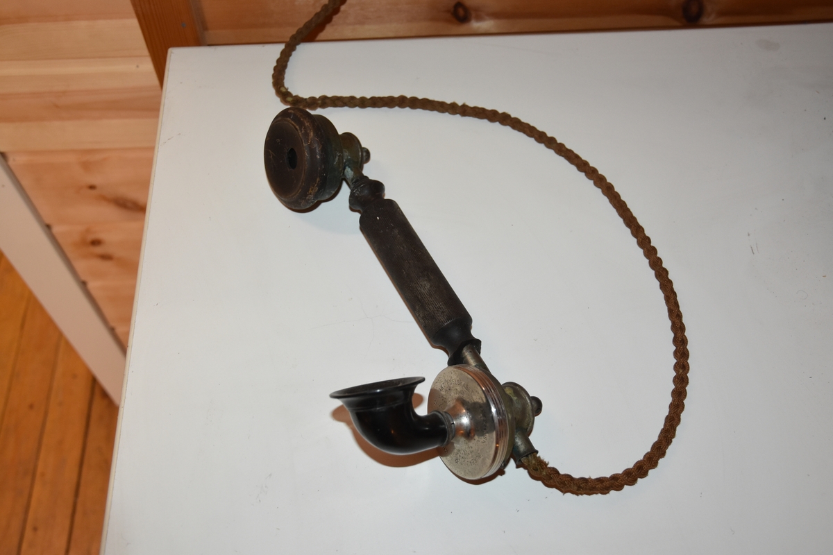 Magneto veggtelefon av blikk med ornamentert støpejern. Treimitasjon og Geometriske dekorasjoner på lokk og fremside som kan minne om treimitasjon. Stoffledning som er tvunnet. Tekst i øvre del. Sveiv på høyre side. Røret med høytaler og"taltetut" henger på venstre side.