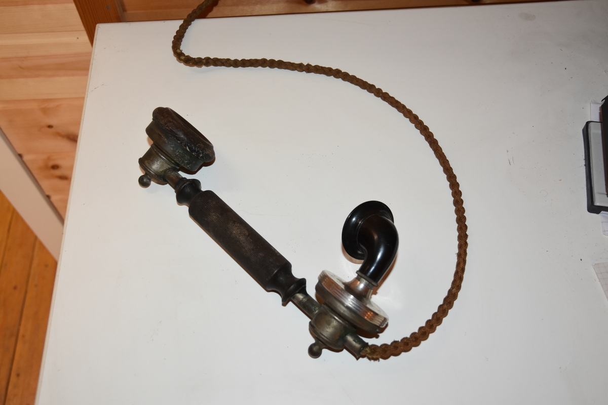 Magneto veggtelefon av blikk med ornamentert støpejern. Treimitasjon og Geometriske dekorasjoner på lokk og fremside som kan minne om treimitasjon. Stoffledning som er tvunnet. Tekst i øvre del. Sveiv på høyre side. Røret med høytaler og"taltetut" henger på venstre side.