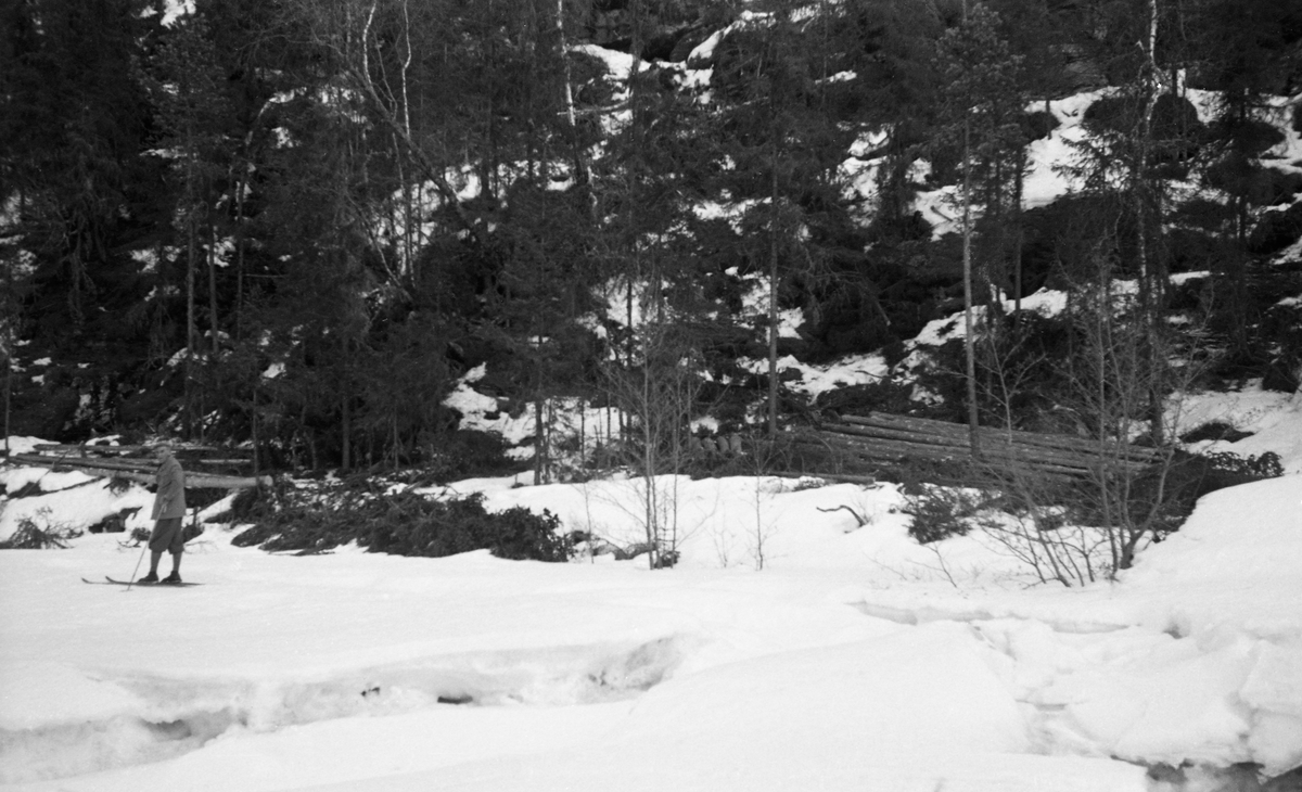 «Tømmertillegging på snøen» ved Vinstra i Nord-Fron i Midt-Gudbrandsdalen. Fotografiet ble tatt i april 1954. Fotografen later til å ha stått på elveisen med kameraet vendt mot elveskråningen, der noen hadde felt og kvistet enkelte grantrær som var samlet i små lunnevelter i skogen langs elvebredden. Det var ikke slik tømmermålerne og fløterne forventet å finne tømmeret. Målerne ville ha det samlet i flaker eller strøvelter, der de kunne inspisere hver enkelt stokk - ikke vasse i snø mellom hver stokk. Og fløterne ville helst ha det i velter på steder der det var enkelt å ruller stokkene ut i elveløpet når elva fikk høvelig vannføring.