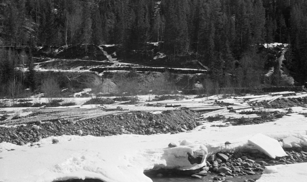 Tømmertillegging ved Gaupvollen, altså i den delen av Vinstravassdraget som renner gjennom Skåbu i Nord-Fron kommune i Midt-Gudbrandsdalen. Dette fotografiet ble tatt i april 1954. I forgrunnen ser vi tvers over et elveløp hvor det fortsatt lå betydelige mengder is, men hvor vannføringa var minimal. Inne på elvebredden lå det barket tømmer i såkalte lunnevelter, der stokkenes lengderetning var parallell med elvas strømretning. Dette tømmeret skulle rulles ut i elveløpet når vannføringa ble stor nok til at Vinstra kunne bære stokkene østover og nedover mot Lågen. At fløtingsfunksjonæren som har tatt dette fotografiet viet elveløpet i dette vassdraget stor oppmerksomhet nettopp i 1954 skyldtes antakelig at det da foregikk store dambarbeider ved Vinstras utløp fra sjøen Olstappen, et prosjekt som kom til å påvirke vannføringa og dermed fløtingsmulighetene i dette elveløpet.