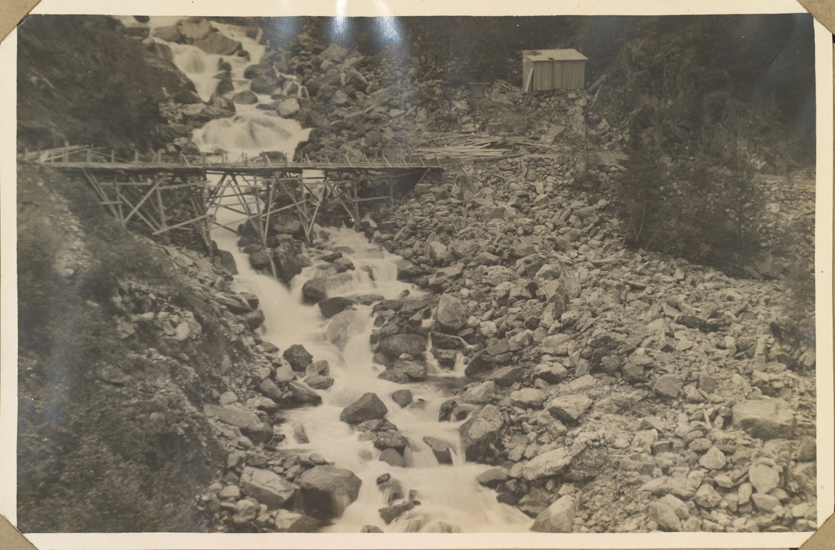 Ødeleggelser etter flommen på Rjukan 28.-29. juni 1927. Ødeleggelser av vei og bro ved Våerbekken. Trebro over elven, lite bygg stående på broens høyre side.