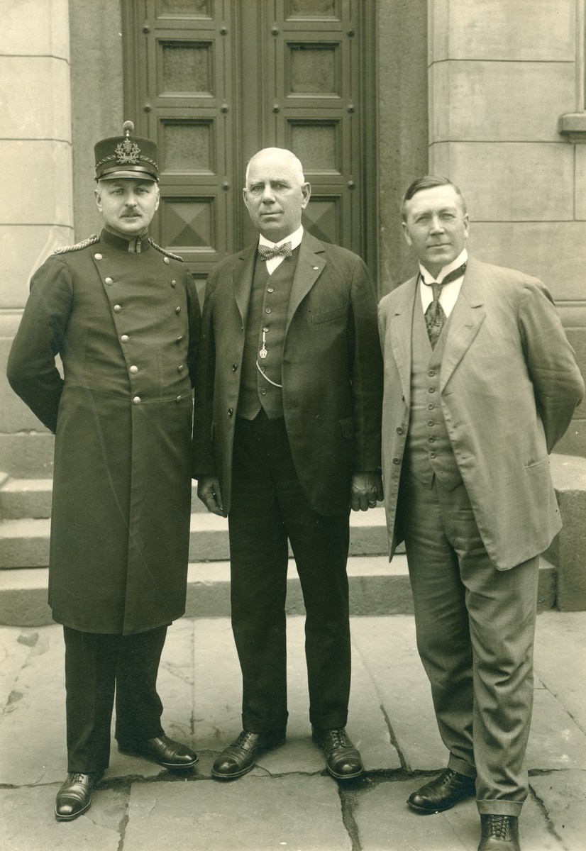 Tre menn foran en dør. En er iført uniform for politiembetsmann modell 1911. De to andre er i sivil.