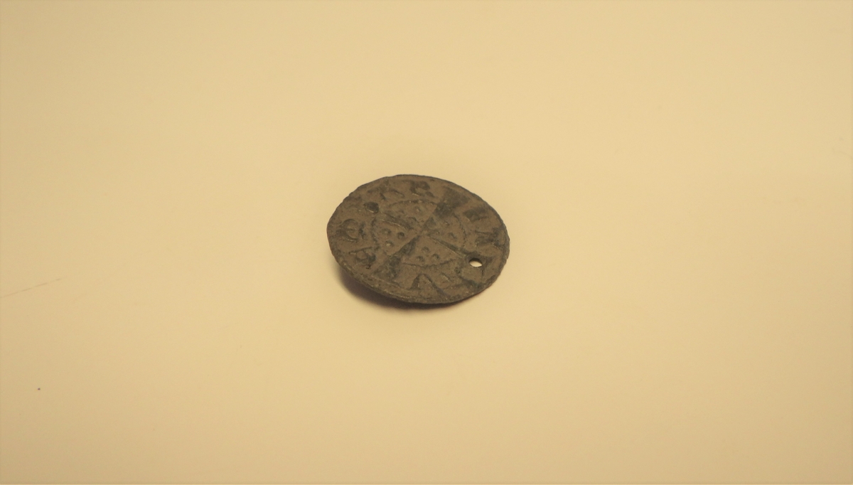 Motivet på gjenstanden kommer fra revers side på de engelske penningene som ble brukt i perioden 1250-1350. Motivet viser et kors som strekker seg over hele siden, og i hvert av korsvinklene sees tre perler. lengre ut  går en linje som former en sirkel  og utenfor denne sees en legende.