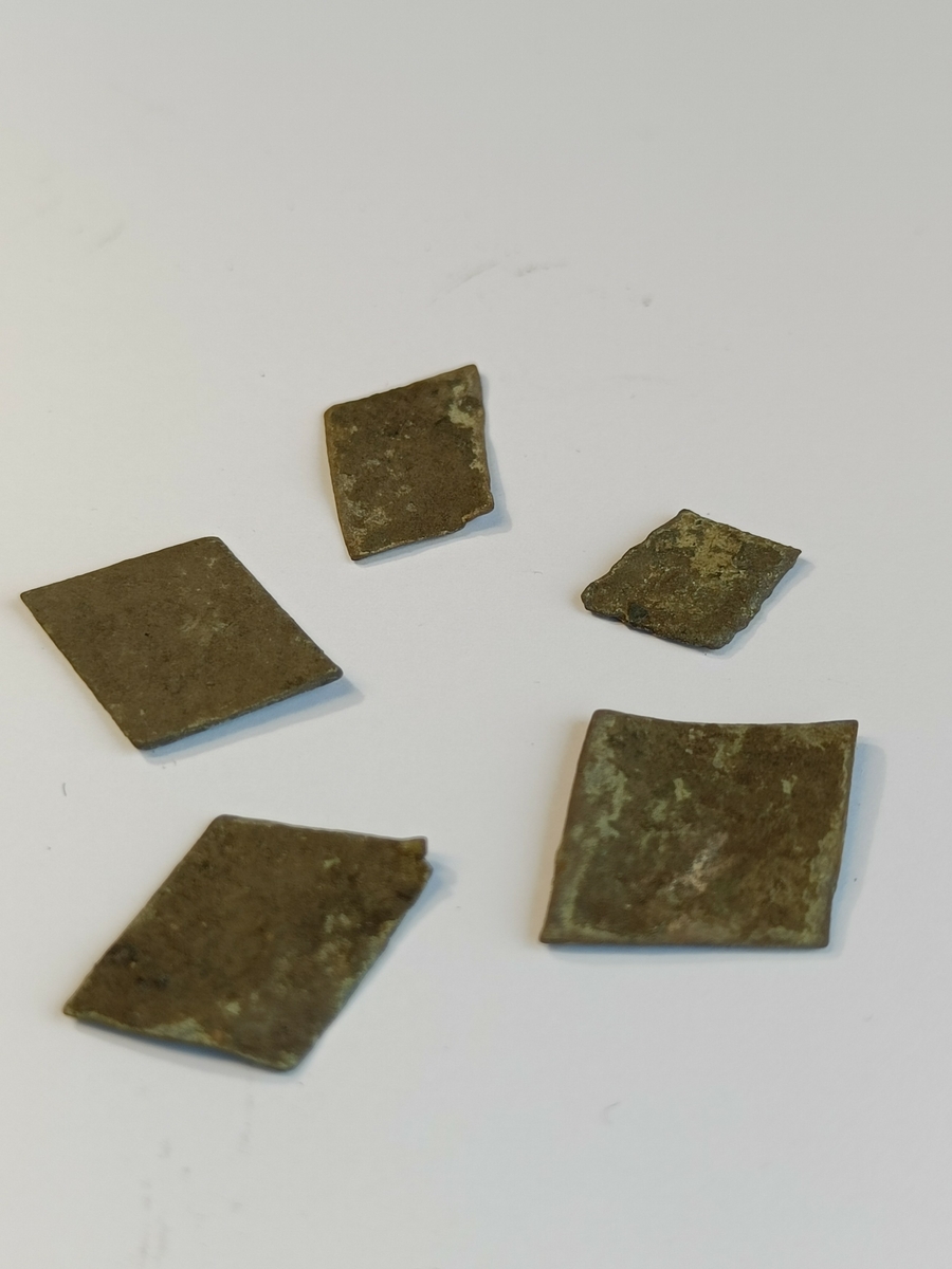 Fem stk små, rombeformede og flate metallplater. Dette er emner til klinknagler.