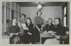 En grupp unga kvinnor har samlats i ett kontor på varvet för fotografering. De hanterar med olika papper för att demonstrerar kontorsarbete.