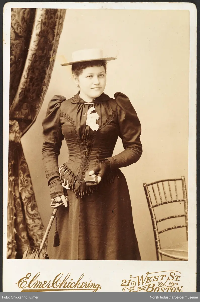 Visittkortfoto, kvinne fotografert i studio. Kvinnen er kledd i kjole med kosettliv, halvfingerhansker og hatt på hodet. Hun holder en paraply i den ene hånden og en lommebok i den andre. Bak henne står det en stol på høyre side og en gardin på venstre side.
