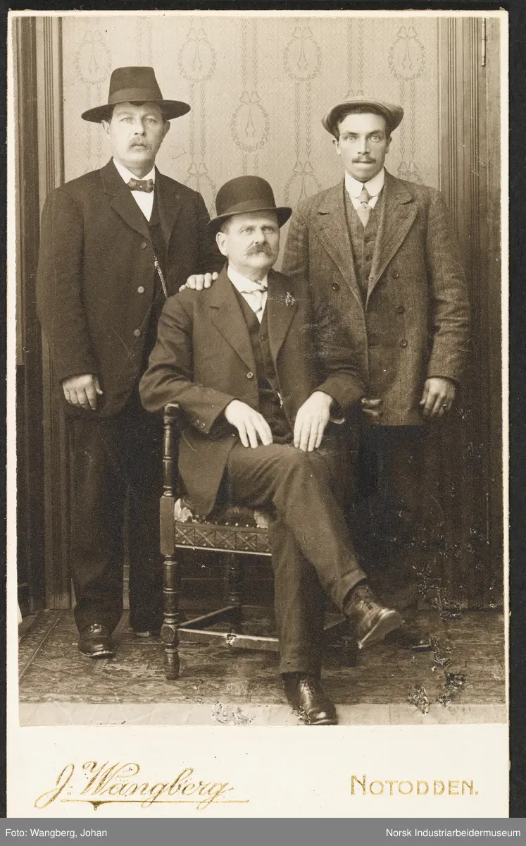 Visittkortfoto, tre menn fotografert i studio. Alle tre er kledd i dress. En av mennene sitter foran de andre i en stol.