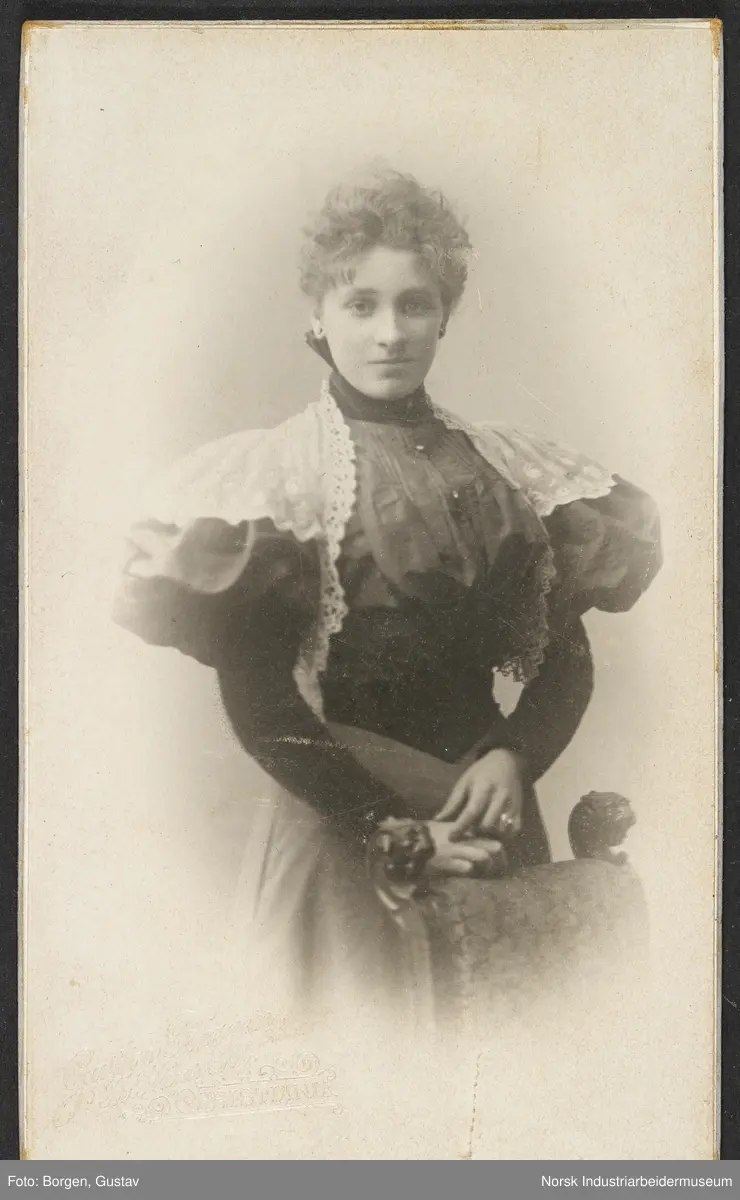 Visittkortfoto, kvinne fotografert i studio. Kvinne kledd i kjole med blondedetaljer og puffede ermer. Stol stående foran henne.