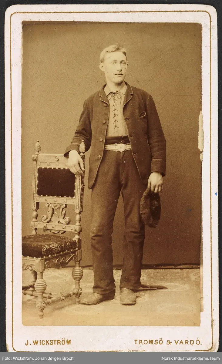 Visittkortfoto, mann fotografert i studio. Mannen står lent mot en stol med fløyelstrekk. Kledd i dress og skjorte med lissesnøring. Holder hatt i venstre hånd.