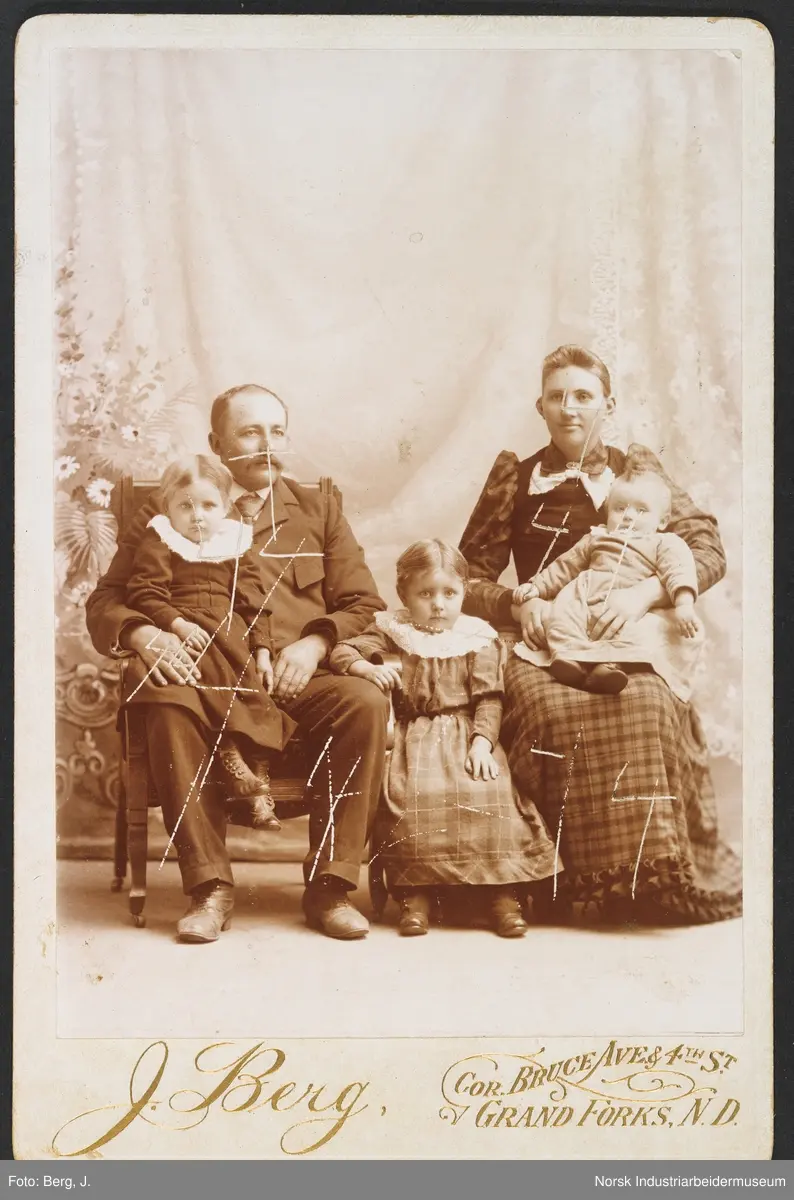 Visittkortfoto, Familie fotografert i studio. Kvinne og mann med tre jenter kledd i pentøy. Den yngste jenta sitter på kvinnens fang, den eldste jenta sitter på stol mellom foreldrene, den mellomste jenta sitter på farens fang.