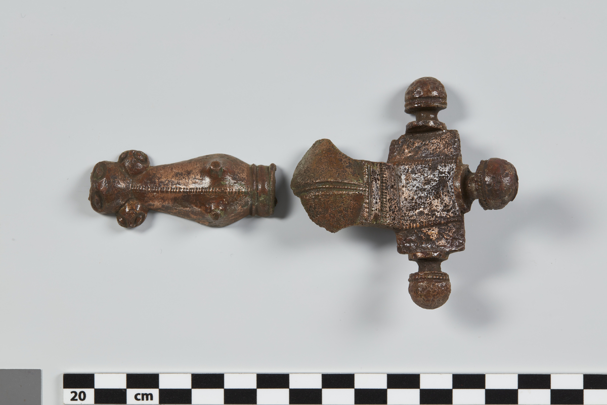 En stor bøileformet Broncespænde af Formen med 3 Knopper øverst og Dyrehoved i nedre Ende (Sv. forns. 326); er overbrudt ved Bøilens nedre Rod, og af begge Stykker mangler den nederste Del. Knopperne flade under, runde ovenpaa; have en halvcirkelformet opstaaende Kam indad mod Midtstykket; Bøilen er stærkt ophøiet i Midten og har Fliger til Siderne; den fortsætter sig som et ophøiet Baand over Midtstykket paa Spændens øvre Parti (cfr. Aarsb. 1875 fig. 21). Dyrehovedets nedre Ende delt i 3 Fliger, saaledes at hver Flig ogsaa har Udseende af et Hoved med Øine. Paa flere Steder prydet med de bekjendte Hak i Kanterne mellem parallele Linier. Paa Bagsiden Spor af en Naal med Spiralsnoning af Jern. Oprindelig Længde vel ca. 15 cm. (5 3/4").