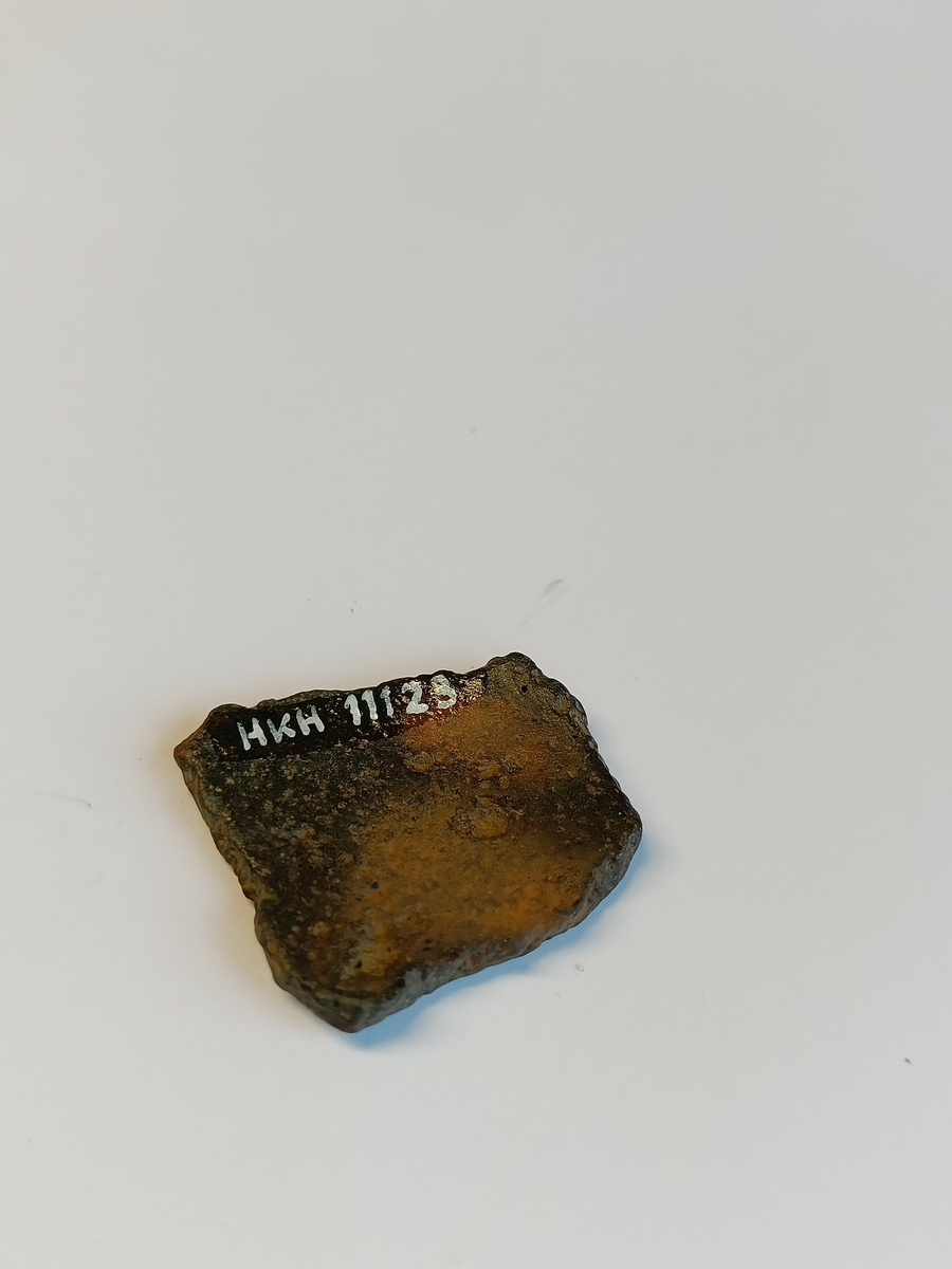 Et svakt buet gryteskår med bruddflater langs alle kanter. Dette er et bukskår fra ei bronsegryte.