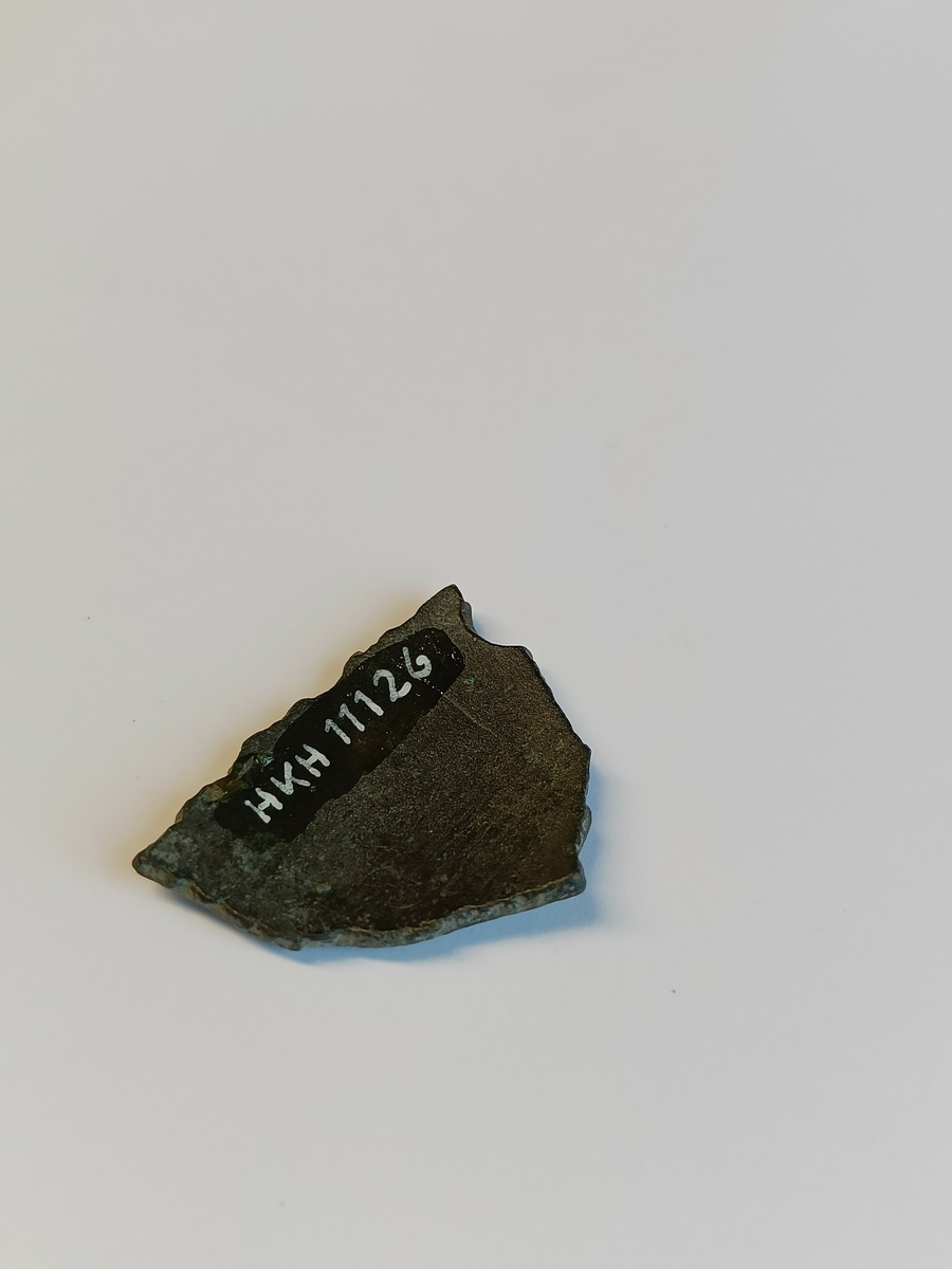 Et lite og trekantet gryteskår med jevn krumming og bruddflater langs alle kanter. Dette er et bukskår fra ei bronsegryte.