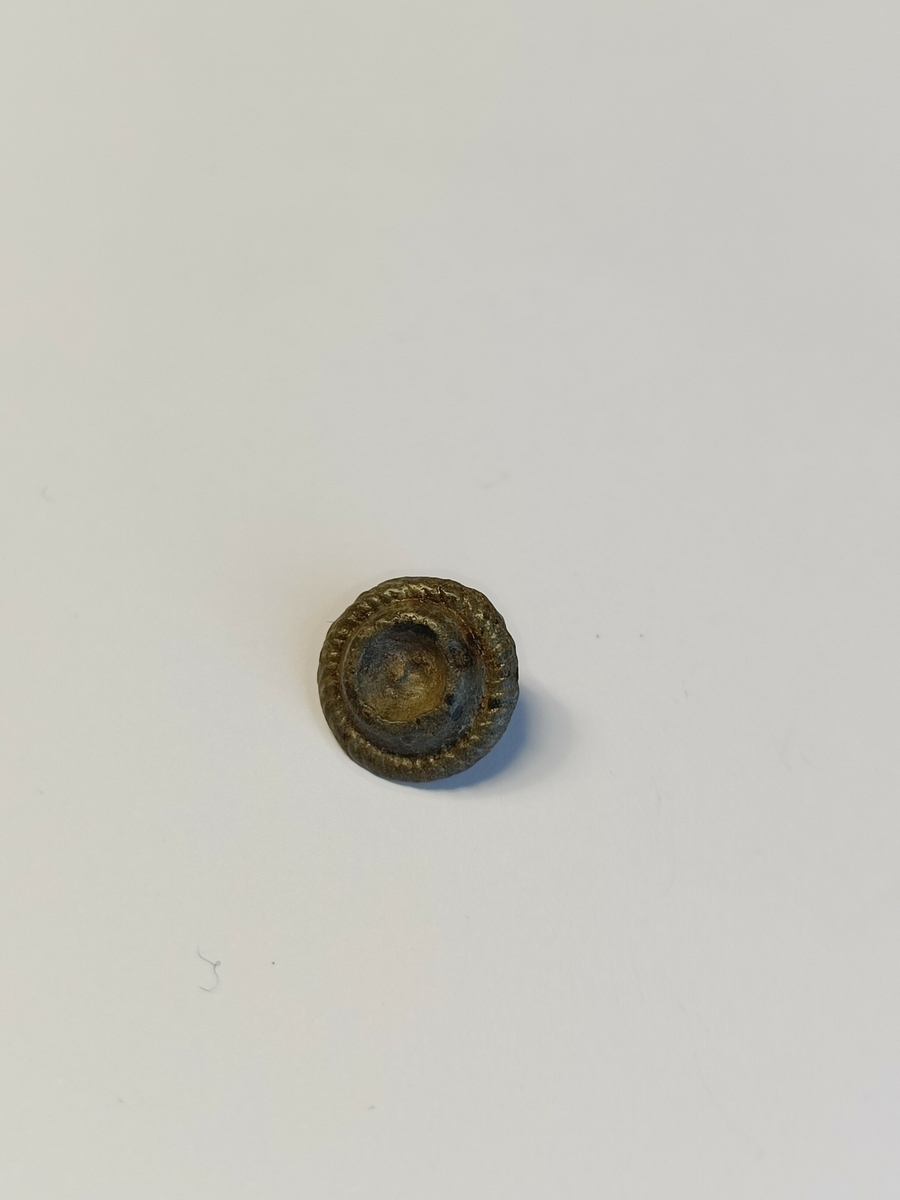 Liten, sirkulær pyntenagle med en enkel nagle på baksiden. Naglen har dekor av et metallbånd med strekdekor langs kanten som imiterer et tvunnet bånd. I midten av naglen sees en forhøyet ring med en fordypning i midten.