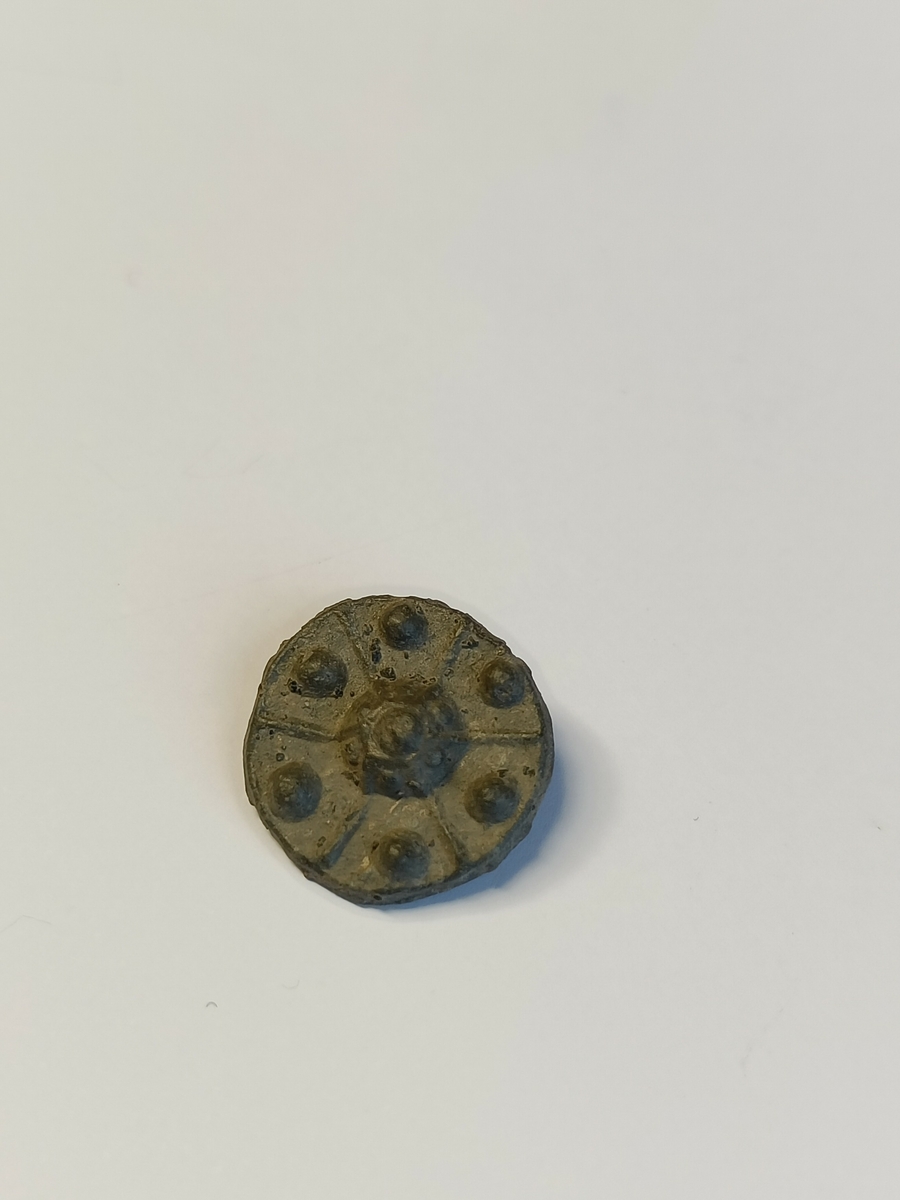 En pyntenagle med rest av en naglestilk på baksiden. Naglehodet er flatt men har opphøyd strekdekor som danner felt ut fra midten, og i hver av disse feltene samt i midten sees opphøyde perler.