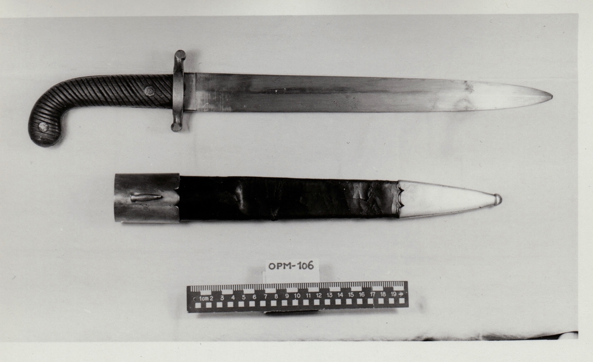 Dolk med rett tveegget klinge. Parerstangen er rett, baktil nedsvunget, fortil oppsvunget, og delt langsetter med en spalte i hver ende. Grep og knapp i ett stykke. Knappen er framsvunget. Skjefteplater i skråriflet tre, festet til gangen med to skruer. Balgen har munnblikk og doppsko, til munnblikket er festet en hempe.