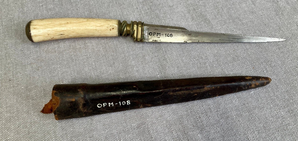 Trekantet, spiss dolk med lærslire. Mellom klinge og grep er det et kroppstykke i vakkert forarbeidet messing. Grepet av elfenben eller hvalrosstann er kronet av en messingknapp med 5 vinkelskårne riller.