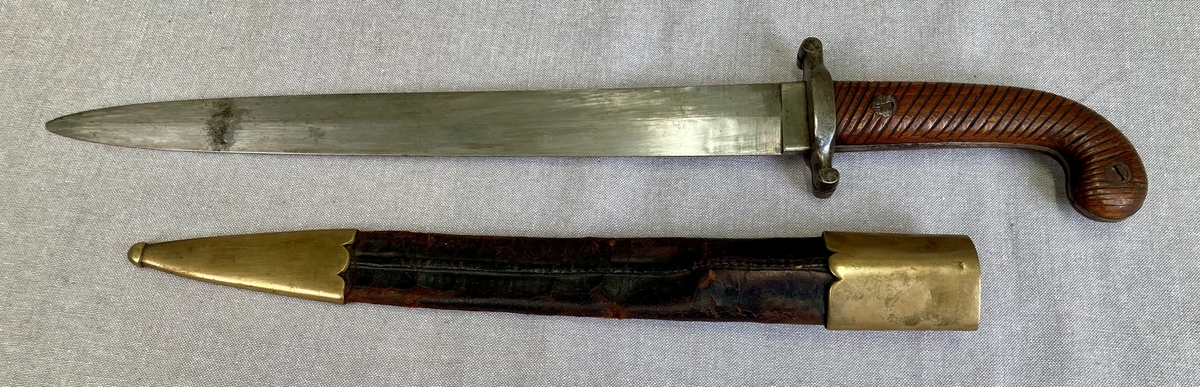 Dolk med rett tveegget klinge. Parerstangen er rett, baktil nedsvunget, fortil oppsvunget, og delt langsetter med en spalte i hver ende. Grep og knapp i ett stykke. Knappen er framsvunget. Skjefteplater i skråriflet tre, festet til gangen med to skruer. Balgen har munnblikk og doppsko, til munnblikket er festet en hempe.