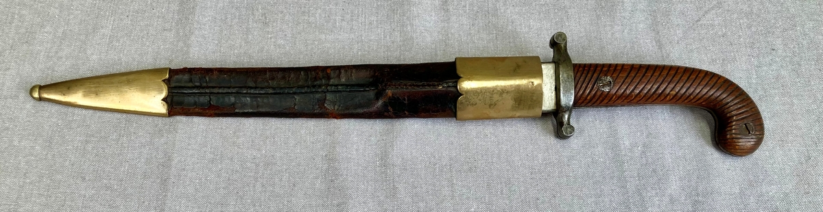 Dolk med rett tveegget klinge. Parerstangen er rett, baktil nedsvunget, fortil oppsvunget, og delt langsetter med en spalte i hver ende. Grep og knapp i ett stykke. Knappen er framsvunget. Skjefteplater i skråriflet tre, festet til gangen med to skruer. Balgen har munnblikk og doppsko, til munnblikket er festet en hempe.