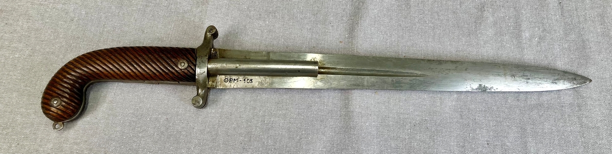 Postførerverge er et et kombinert stikk- og skytevåpen. 

Klingen er rett, og har hulkiler (blodrenner) på hver side. Grep og knapp er i ett stykke, knappen er svunget fremad, grep og knapp har skjefteplater av tre med skråttstilte riller og er festet til tangen med to nittebolter. Fra parerstangen går det på hver side av klingen et 10 mm pistolløp, de hviler i hulkilene. Pipene er skrudd fast til parerstangen, som er delt på langs, det to bakre deler utgjør slaghaner, som ved å spennes blottlegger to pistonger. Når hanene spennes, felles to avtrekkere ut på framsiden av grepet. 

Balgen har munnblikk og doppsko av messing, på minnblikket en messinghempe. Klingen er merket med en kronet K. Parerstangen er merket OL 1848. Hanen er merket med produksjonsnummer 12.