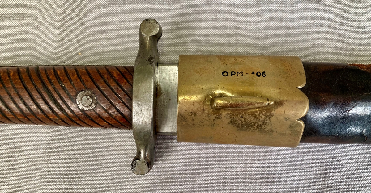 Dolk med rett tveegget klinge. Parerstangen er rett, baktil nedsvunget, fortil oppsvunget, og delt langsetter med en spalte i hver ende. Grep og knapp i ett stykke. Knappen er framsvunget. Skjefteplater i skråriflet tre, festet til gangen med to skruer. Balgen har munnblikk og doppsko, til munnblikket er festet en hempe.