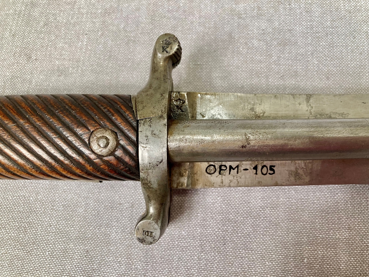 Postførerverge er et et kombinert stikk- og skytevåpen. 

Klingen er rett, og har hulkiler (blodrenner) på hver side. Grep og knapp er i ett stykke, knappen er svunget fremad, grep og knapp har skjefteplater av tre med skråttstilte riller og er festet til tangen med to nittebolter. Fra parerstangen går det på hver side av klingen et 10 mm pistolløp, de hviler i hulkilene. Pipene er skrudd fast til parerstangen, som er delt på langs, det to bakre deler utgjør slaghaner, som ved å spennes blottlegger to pistonger. Når hanene spennes, felles to avtrekkere ut på framsiden av grepet. 

Balgen har munnblikk og doppsko av messing, på minnblikket en messinghempe. Klingen er merket med en kronet K. Parerstangen er merket OL 1848. Hanen er merket med produksjonsnummer 12.