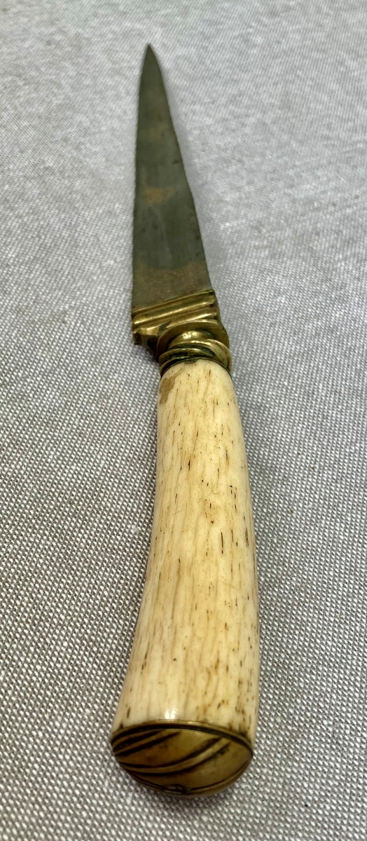 Trekantet, spiss dolk med lærslire. Mellom klinge og grep er det et kroppstykke i vakkert forarbeidet messing. Grepet av elfenben eller hvalrosstann er kronet av en messingknapp med 5 vinkelskårne riller.