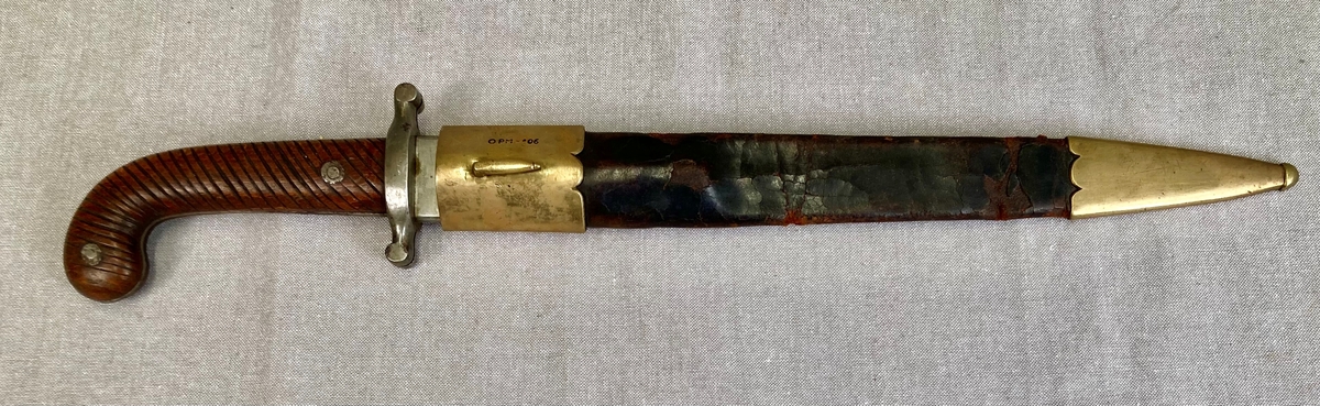 Dolk med rett tveegget klinge. Parerstangen er rett, baktil nedsvunget, fortil oppsvunget, og delt langsetter med en spalte i hver ende. Grep og knapp i ett stykke. Knappen er framsvunget. Skjefteplater i skråriflet tre, festet til gangen med to skruer. Balgen har munnblikk og doppsko, til munnblikket er festet en hempe.