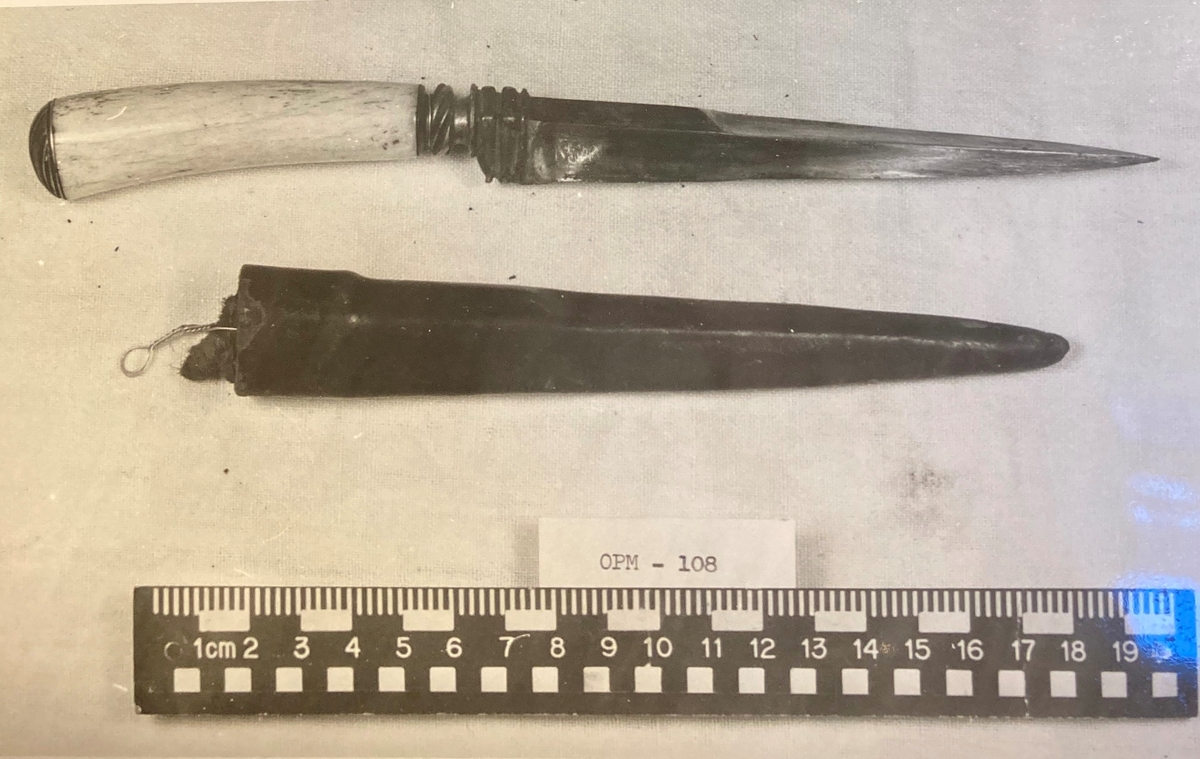 Trekantet, spiss dolk med lærslire. Mellom klinge og grep er det et kroppstykke i vakkert forarbeidet messing. Grepet av elfenben eller hvalrosstann er kronet av en messingknapp med 5 vinkelskårne riller.