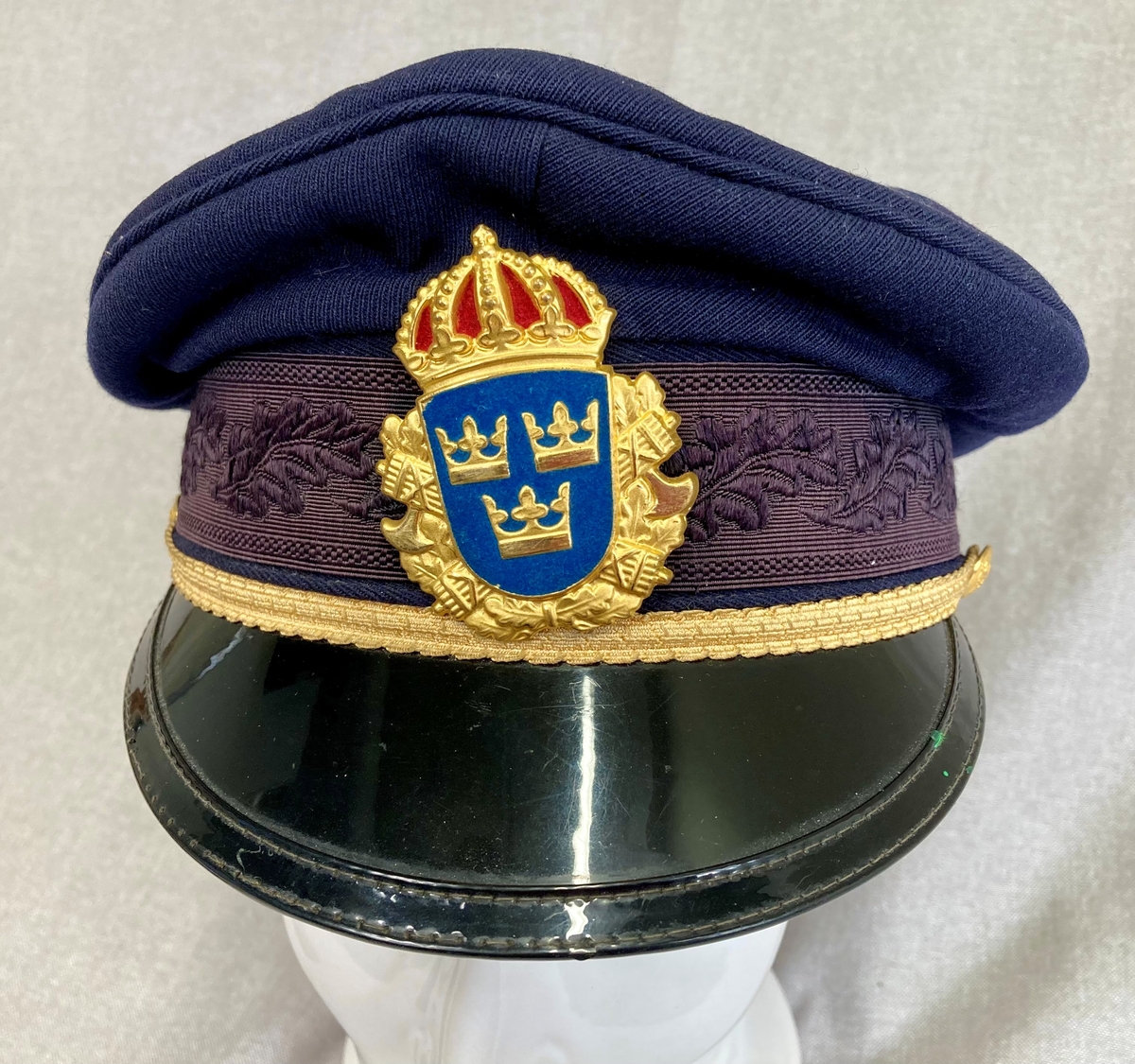 Uniformslue for svensk politi. Mørkeblå med gulldetaljer og svensk politimerking. Luebånd med eikebladsdekor. Blank sort skjerm.