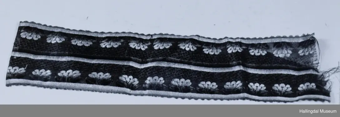 Rød bunn. I midten en rosa og en grønn stipe. Langs ene siden rosa striper, langs denne gule blomster og grønne blad. Langs andre siden gul stripe, rosa blomster og grønne blad. I ene enden er 3,2 cm brettet inn og tråklet til. Lå inni HB 01103.