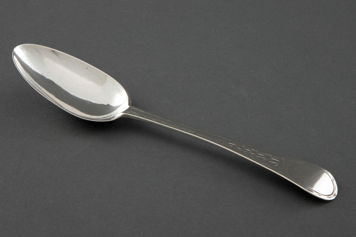 Sølvskje med smalt og ovalt blad. Flatt skaft med tiltagende bredde. Avrundet og noe nedbøyet skaftende. På skaftendenes overside er det gravert et medaljelignende ovalt felt. Skaftroten ender bare tilsynelatende litt inn på skjebladet. Øverst på skaftendenens underside er det stemplet en blomst - Trondheims bymerke. Under dette er det stempla initialer og årstall.