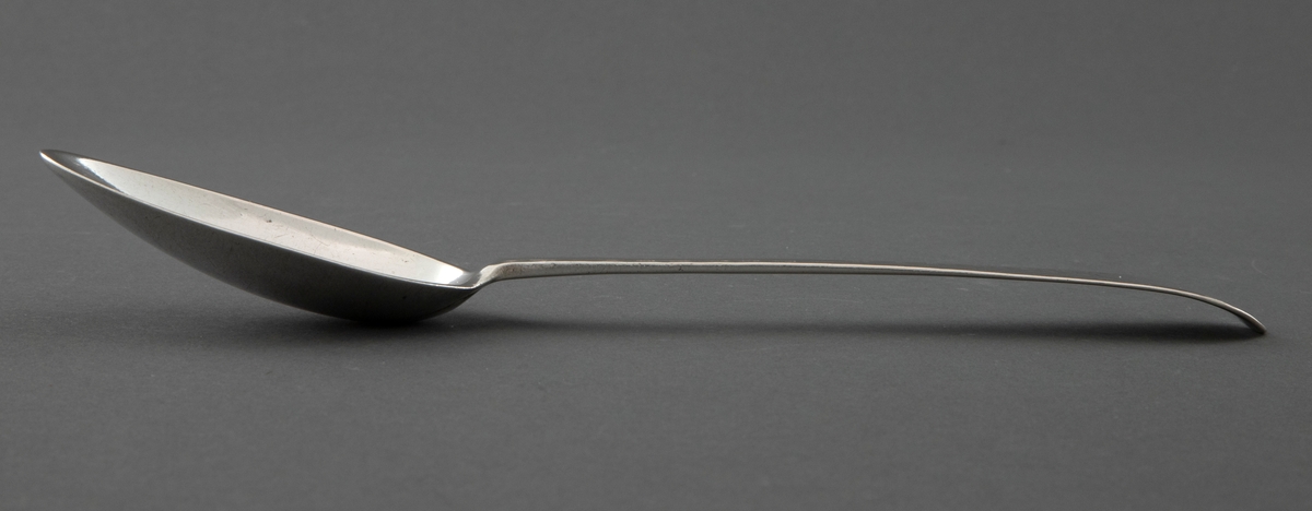 Sølvskje med smalt og ovalt blad. Flatt skaft med tiltagende bredde. Avrundet og noe nedbøyet skaftende. På skaftendenes overside er det gravert et medaljelignende ovalt felt. Skaftroten ender bare tilsynelatende litt inn på skjebladet. Øverst på skaftendenens underside er det stemplet en blomst - Trondheims bymerke. Under dette er det stempla initialer og årstall.