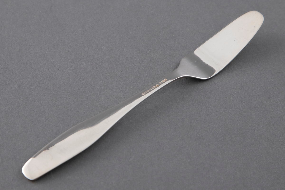 Smørkniv i sølv i ett stykke. Flat skaft som øker ganske sterkt i bredde og minsker lett mot skaftenden. Avrundet og lett oppbøyd skaftende. Flatt, forsenket blad, avsmalnende med rund avslutning.