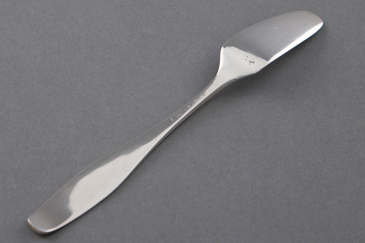 Smørkniv i sølv i ett stykke. Flat skaft som øker ganske sterkt i bredde og minsker lett mot skaftenden. Avrundet og lett oppbøyd skaftende. Flatt, forsenket blad, avsmalnende med rund avslutning.