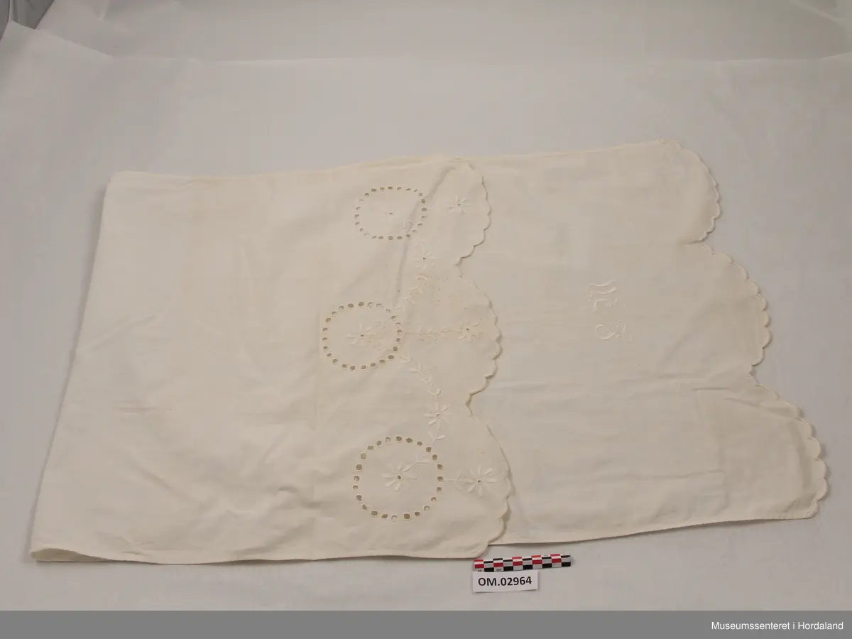 Hvit pyntehåndduk med initialene LM brodert nederst. Øverst er tre ringer formet av sydde hull og brodert blomsterguirlander.