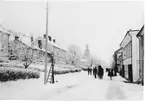 Brahegatan i Gränna vintertid med snön. En liten folksamling kring en häst med släde mitt på gatan. En stege står rest mot en lykta till vänster.