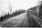 En buss kommer körande på en grusväg vid Stava i riktning mot fotografen. Till höger en gärsgård och två små hus.