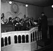 En kör står i en kyrka och sjunger. Några spelar gitarr. En man står och dirigerar kören.