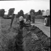 Två män står på en traktor. De dikar en lerväg. En man sitter på grävskopan som gräver ut diket. I utkanten av bilden skymtas ett lastbilsflak.