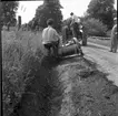 En man tittar på när en traktor dikar en lerväg. En man sitter på grävskopan som gräver ut diket.