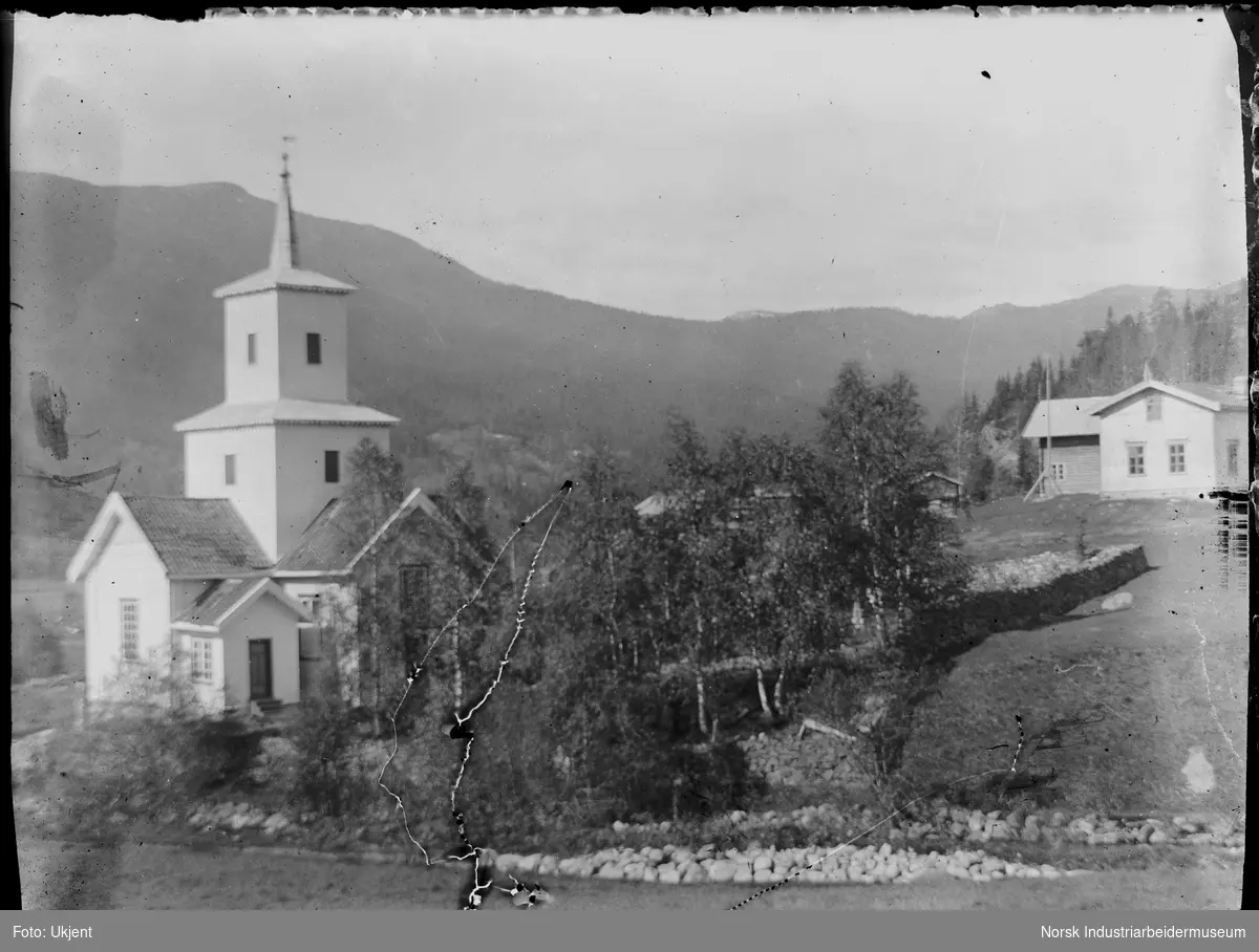 Tuddal kirke og kirkegård. Steingjerde rundt kirkens område, klynge med bjørketrær innenfor steingjerde. Bolighus  med stabbur i bakgrunnen.