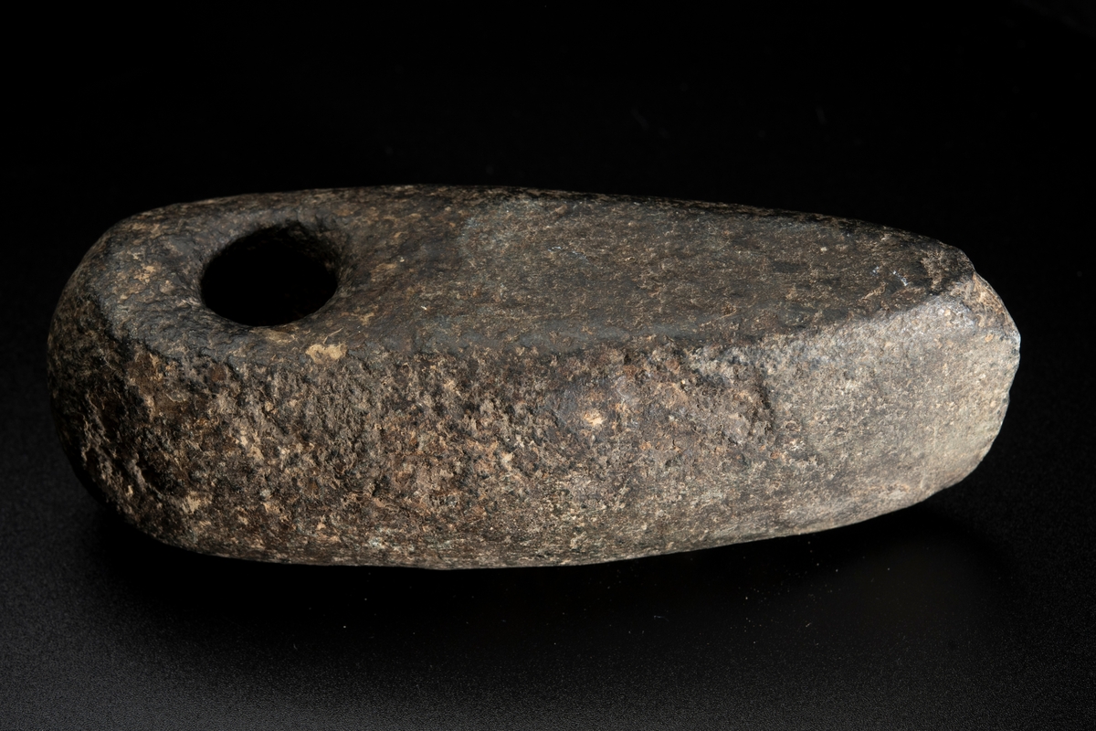 Skafthålsyxa av bergart. Skafthålet placerat långt bakåt. Välvd ovansida, plan undersida. Rundad nacke.