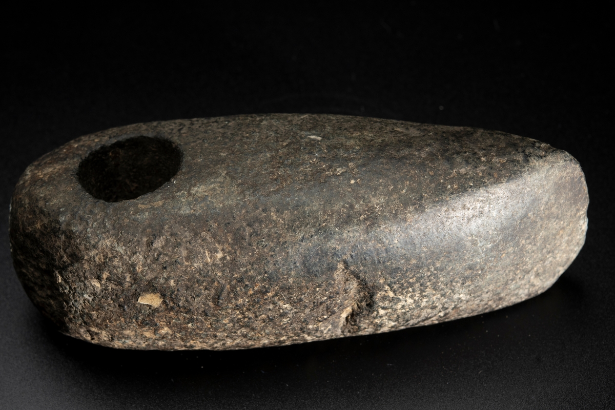 Skafthålsyxa av bergart. Skafthålet placerat långt bakåt. Välvd ovansida, plan undersida. Rundad nacke.