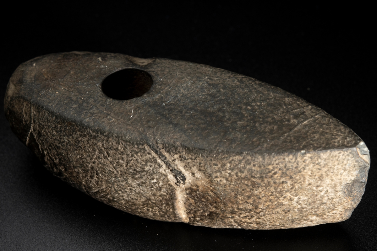 Skafthålsyxa av bergart. Plan ovan- och undersida. Slipad. Yttersta nackpartiet sluttar skarpt.