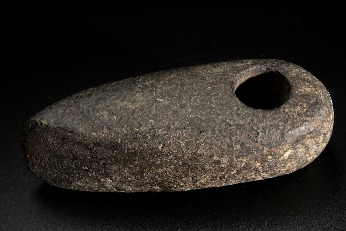 Skafthålsyxa av bergart. Skafthålet placerat långt bakåt. Välvd ovansida, plan undersida. Rundad nacke.