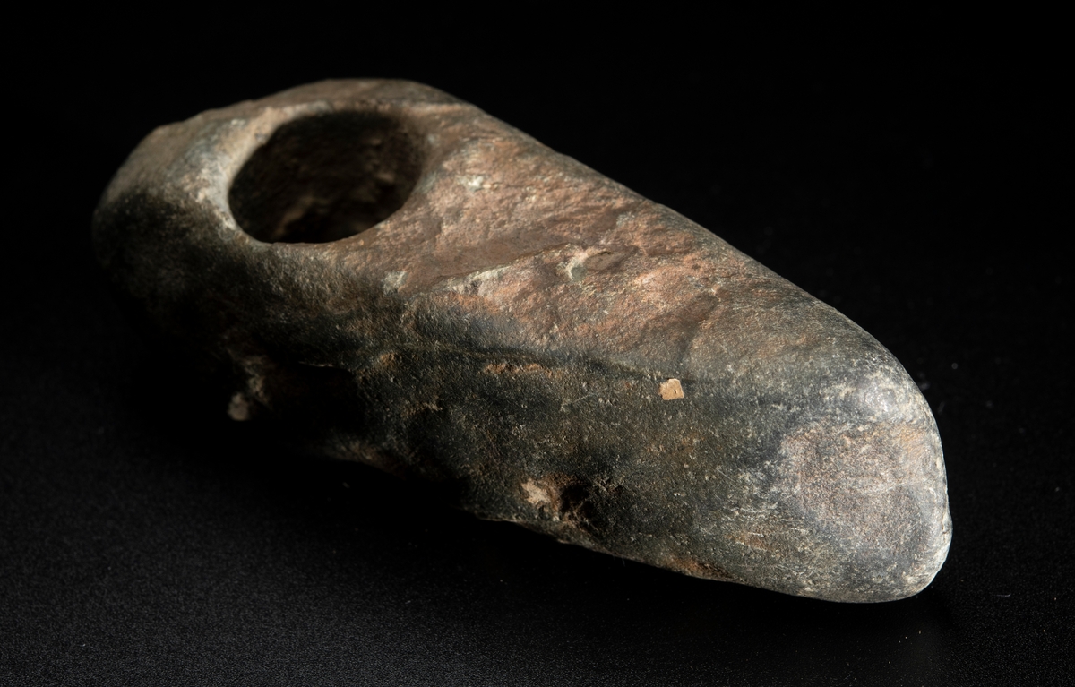 Skafthålsyxa av bergart. Bredast i nacken. Skafthålet placerat långt bakåt. Plan ovan- och undersida.