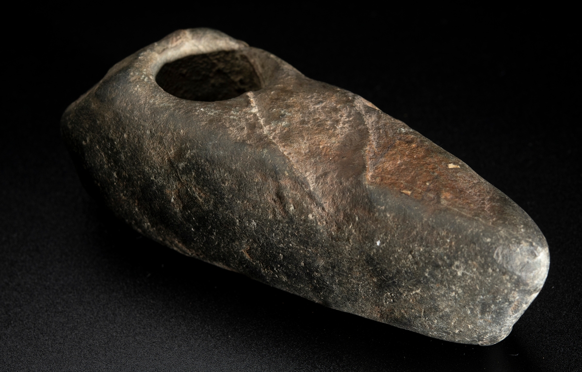 Skafthålsyxa av bergart. Bredast i nacken. Skafthålet placerat långt bakåt. Plan ovan- och undersida.